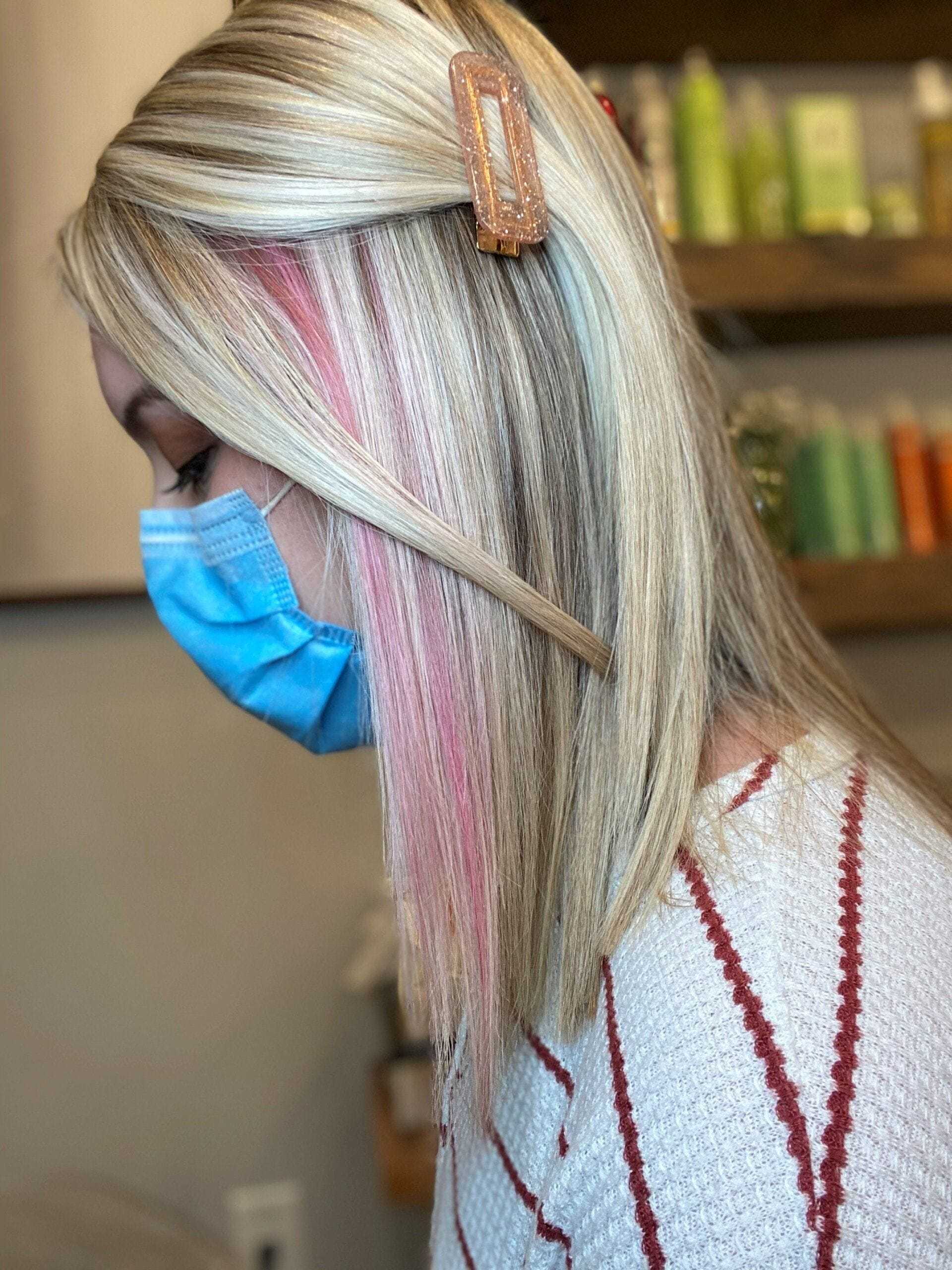 Woman wearing a blue mask with straight, blonde hair and pink highlights, featuring a hair clip.
