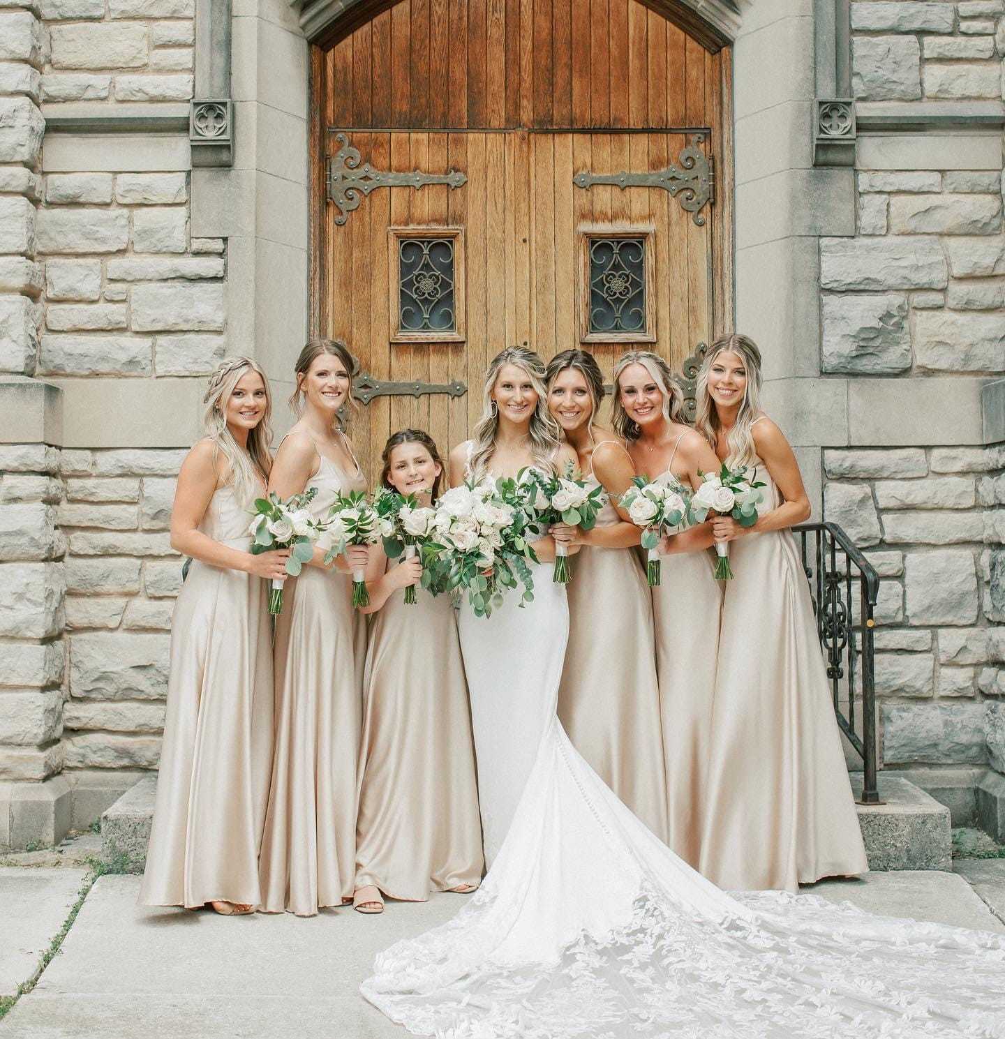 Bride and bridesmaids in beige dresses holding flowers, standing in front of a wooden door.