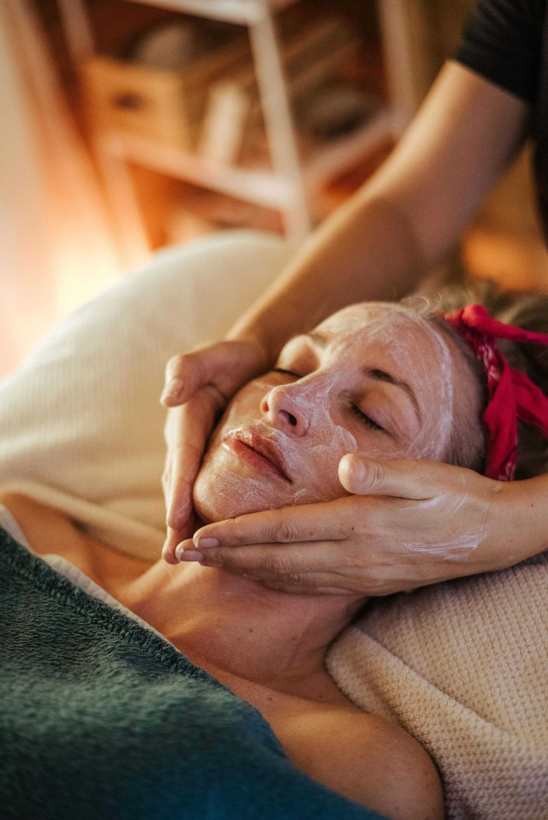 Person receiving a facial treatment with hands applying cream, relaxed on a spa bed.