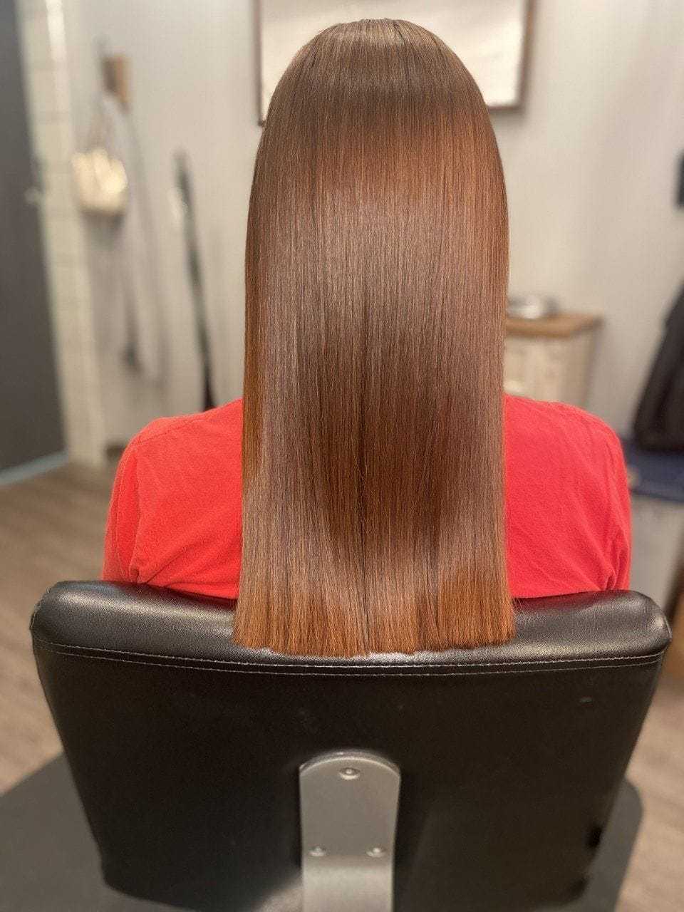 Woman with long, straight, shiny brown hair sitting in a salon chair, wearing a red shirt.